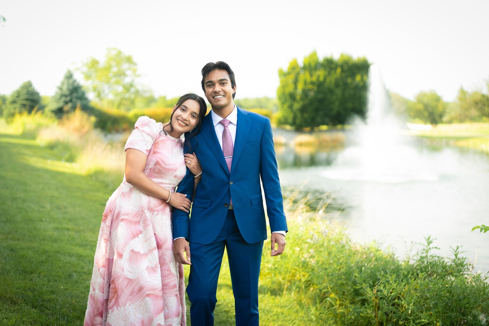 Riyana resting her head on Aadesh's shoulder beside a lake, a fountain behind them